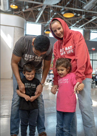 medium-skinned person wearing a grey crewneck t-shirt with best day of my life written looking down at young child wearing a black crewneck with best day of my life. next to them is a light-skinned person wearing a red pullover hoodie with best day of my life standing behind a young child wearing a pink crewneck t-shirt with best day of my life written on it looking at the floor